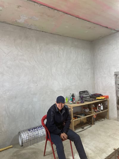 A male individual is seated on a red metal folding chair in an indoor space, likely a workshop, located in the city of Balykchi, Uzbekistan. The subject, wearing a dark blue baseball cap, a dark blue jacket with darker paneling, and dark blue trousers, is positioned slightly off-center to the left, facing the camera with a closed-mouth smile and hands clasped in his lap. The room features light gray walls with a textured, unfinished plaster finish exhibiting swirling patterns. The ceiling is an unfinished light pinkish-beige with visible rough plaster and some peeling, bordered by a thin dark line where it meets the walls. The floor is an unpainted concrete slab. To the left of the subject, a horizontally oriented roll of silver insulation material, bearing the text "EVROIZOL" and "ТЕПЛОИЗОЛЯЦИЯ" in Cyrillic, rests on the floor. To the right, a wooden workbench with two shelves is positioned against the wall, laden with various items including a black toolbox containing a socket set, a purple power drill, multiple plastic bottles, and orange and yellow plastic containers. Beneath the workbench are cardboard boxes and a white bucket. A metal storage rack is partially visible on the extreme right of the frame.