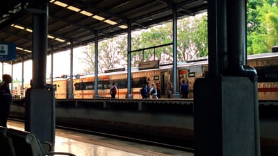 The image depicts a train station platform in Karawang, Indonesia. Several individuals are waiting near a stationary train. Overhead signage indicates the location as "Karawang." The station has a metal roof supported by pillars. The environment includes greenery visible beyond the platform. The train has a silver exterior with red stripes, and open doors suggest boarding or disembarking. The lighting suggests early morning or late afternoon.