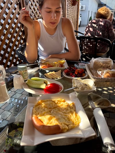 The image depicts a table set for breakfast outdoors, likely at a cafe or restaurant in Parakar, Armenia, given the context provided. A young woman, wearing a light blue tank top and a silver necklace, is seated at the table, holding a fork and looking down at her food. Sunlight streams in from the right, casting shadows and highlighting the scene.
On the table, several plates of food are arranged. One plate contains scrambled eggs and a sausage. Another holds sliced cucumber, tomato, and a small dish of what appears to be jam. A third plate features sliced cheese and a cluster of cherries. A basket filled with bread, possibly croissants or rolls, is also visible. A small white dish contains a creamy spread, with a spoon resting in it.
To the left, there is a salt shaker, a glass container, and a small green and grey tube labeled "TOOTHpicks." A menu with the word "MENU" prominently displayed and some Armenian text is also on the table. In the background, part of the outdoor seating area is visible, including a wooden lattice fence and a table with other diners. The overall impression is one of a relaxed morning meal.