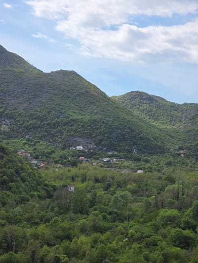 The image depicts a mountainous landscape in Seoca, Montenegro, viewed from a distance. The mountains are covered in lush green vegetation, including trees and other foliage. Several small houses and buildings are nestled amongst the trees on the mountainside. The sky is blue with scattered white clouds. No people are visible in the image.
