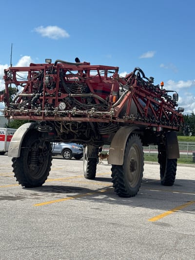 This is a full-shot, low-angle, outdoor photograph of a large, red agricultural sprayer vehicle parked in a paved lot with yellow parking lines. The vehicle has a complex structure of red metal framing, a large liquid tank, and numerous hoses and nozzles. It has four large, off-road tires. A circular sign with "30 km/h" is visible on the side of the vehicle. In the background, there is a U-Haul truck, a silver SUV, a fence, trees, and a blue sky with scattered white clouds. A dog is partially visible in the lower right quadrant of the image, behind the vehicle's front tire. The scene is devoid of any identifiable city landmarks of Syracuse, United States of America.