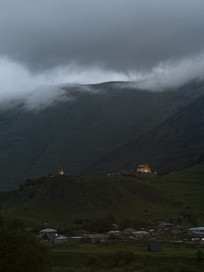 The image depicts a remote village nestled in a mountainous landscape, likely in Sioni, Georgia. The scene is captured at dusk or in twilight, as indicated by the dim lighting and the illuminated windows of several structures. A thick bank of fog or clouds cascades down a steep mountainside on the left, obscuring part of the terrain and creating a dramatic, ethereal atmosphere.

The village itself consists of numerous small houses and buildings clustered together on the grassy slopes. Many of the houses have visible roofs, and some show lights emanating from within, suggesting habitation. The architecture appears rustic and traditional, characteristic of rural mountain communities. The natural setting is characterized by steep, verdant hillsides covered in grass, with a stark contrast between the dark, mist-shrouded slopes and the more brightly lit village.

The overall mood is one of quiet solitude and remoteness. The fog adds a sense of mystery and isolation to the scene. The activity taking place is simply the existence of the village at the end of the day, with life continuing inside the illuminated homes. There are no visible people or explicit actions, but the lights imply human presence. Notable details include the dramatic fog, the scattered lights in the village, and the steep, natural terrain. No text is visible in the image.