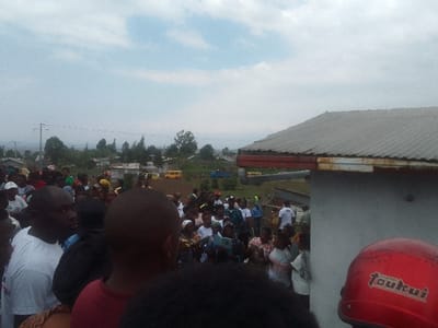 A crowd of people gathers outdoors under a cloudy sky. A corrugated metal roof borders the right side of the frame. In the foreground, a red helmet with white lettering partially obscures the view. Beyond the crowd, a landscape reveals houses and trees, with mountains visible in the distance. A yellow school bus is parked amidst the buildings. The scene suggests a community gathering in a rural or semi-rural setting.