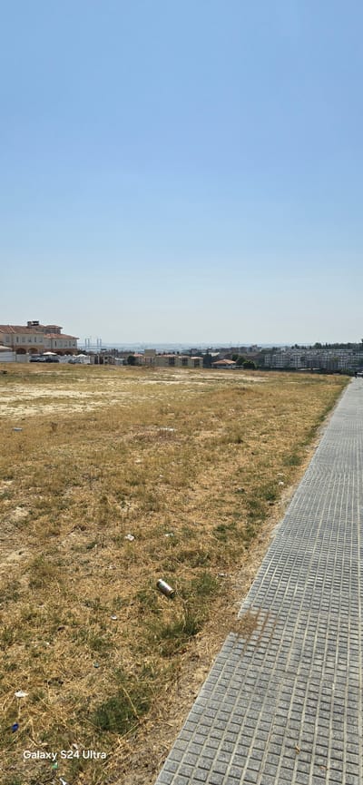 The image shows an open, dry, grassy field with scattered litter, including a can lying on the ground. To the right, a gray tiled sidewalk runs along the edge of the field. In the background, a few buildings are visible, including a larger building with a reddish-brown roof on the left and several residential buildings. In the far distance, a clear sky is visible, with hints of structures and possibly a bridge. The overall impression is that of an undeveloped or partially developed area within a city. The text "Galaxy S24 Ultra" is overlaid in the bottom left corner of the image. The scene appears to be in San Juan de Aznalfarache, Spain.