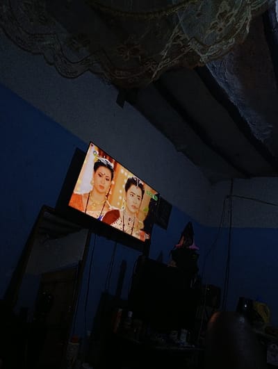 An indoor scene in a dimly lit room, identified as Vanga ward, Kenya. The room features dark blue walls and a ceiling with exposed dark beams from which light-colored, possibly lace, patterned fabric is draped. A flat-screen television, mounted or placed on a stand, occupies the center of the image, actively displaying content. The screen shows two women in traditional South Asian attire and elaborate jewelry, consistent with a dramatic program. The woman on the left wears gold and red with gold jewelry, while the woman on the right wears maroon with silver-toned jewelry and a bindi. The channel logo "Swadhn" is visible on the screen. Below the television, a dark, rectangular object, likely another electronic device or piece of furniture, is present. To the left of the television, a tall, dark reflective surface, possibly a mirror or dark window, is visible. Various indistinct objects, appearing as clutter, are present on surfaces in the foreground. A blurry, dark shape, consistent with a person's shoulder or head, occupies the lower right foreground. The primary illumination in the room emanates from the television screen, casting deep shadows throughout the environment.