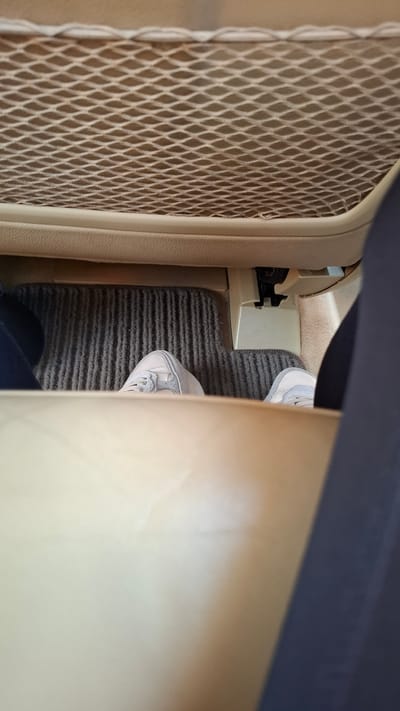 The image shows a person's feet, wearing light-colored sneakers, resting on a grey carpeted floor inside a vehicle. The feet are positioned in the footwell, with a beige mesh pocket visible above. The interior of the vehicle appears to be beige and dark grey. The image was taken in the city of Malo Buchino, Bulgaria.
