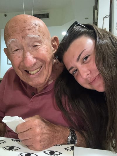 The scene is set indoors, presumably within a dining or home environment, located in Bnei Dror, Israel. The image features two individuals positioned closely together, from the chest up.

On the left, an elderly male individual with a bald head, white eyebrows, and visible facial wrinkles exhibits a wide, open-mouthed smile, showing his teeth. He wears a reddish-brown collared polo shirt. His right hand, positioned in the lower-left portion of the frame, holds a crumpled white napkin or tissue. A metallic wristwatch with a black face is visible on his left wrist.

On the right, a younger female individual with long, dark hair leans her head against the male's right shoulder. She has black sunglasses resting on top of her head and a small stud piercing visible on her left nostril. She displays a subtle smile, with her eyes directed towards the camera. Her attire is a dark-colored top.

The foreground includes a table covered with a white and black textile featuring a repeating pattern of stylized eyes. The background consists of a light-colored wall, a rectangular air vent mounted high, and a framed picture partially visible on the left side of the wall. The scene is illuminated by overhead lighting.