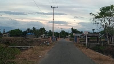 The image depicts a straight, paved road leading into the distance in Pasi, Leubeue, Indonesia. The road is flanked by dry, grassy embankments on either side. Small, simple houses with tin or tile roofs are visible on the left side of the road, surrounded by trees. On the right side, there are more trees and some open-sided structures. Several utility poles with wires are spaced along the road, indicating the presence of electricity. The sky is overcast with a mix of gray and lighter clouds, suggesting it is either early morning or late afternoon. The overall atmosphere is rural and somewhat undeveloped. There are no people or animals visible in the scene.