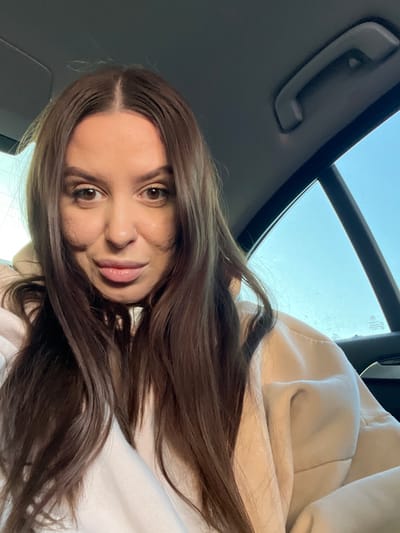 A young adult female is depicted in a close-up, chest-up view, seated within the rear passenger compartment of a vehicle in the city of Svinoedovo, Russia. She has long, straight, dark brown hair with a middle part, dark brown eyes, and full lips, looking directly into the camera with a neutral expression. Her complexion is fair, exhibiting some textural variations on her cheeks. She is dressed in a light-colored undershirt, possibly white, layered beneath a beige hooded garment. The car's interior is dark, featuring a gray grab handle on the ceiling and a visible window frame. A clear, light blue sky is visible through the car window on the right, which also displays a faint BMW logo. The scene is illuminated by natural daylight.