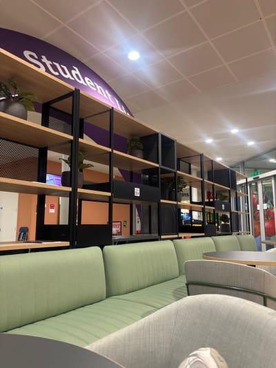 An interior space showcases shelving units with black frames, interspersed with plants and closed cabinets in various colors. A long, green upholstered bench with vertical stitching sits parallel to the shelves. Adjacent, there are light gray upholstered chairs and tables. The ceiling is bright with multiple lights, and a section of the shelf is labeled with "Student." The floor appears to be a light color.