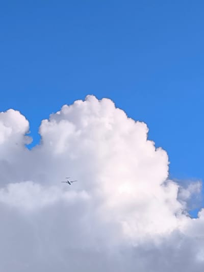 An airplane is captured in mid-flight against a bright blue sky, partially obscured by a large, fluffy white cloud. The airplane appears to be a turboprop aircraft, with its twin engines and propeller visible. It is flying from left to right across the frame. The cloud, a cumulus formation, dominates the lower half of the image, with its white, textured surface catching the sunlight. The sky is a clear, vibrant blue, offering a stark contrast to the white of the cloud and the gray of the lower cloud edges. There are no discernible people or structures related to the city of Vētras, Latvia, visible in this image.