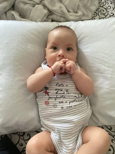 This media file is a close-up, overhead shot of a baby lying on a white pillow. The baby is wearing a white and navy striped onesie with nautical-themed designs, including anchors and ship wheels. There is red lettering on the onesie, but it is partially obscured by the baby's arm. The baby has its hands near its mouth and is looking towards the camera with wide eyes, appearing curious and alert. A thin red string is visible on the baby's left wrist. The setting appears to be indoors, likely a bed, with a white blanket to the left and a decorative black and white patterned fabric to the far left and right. The lighting is soft and natural, suggesting daytime. The baby's facial expression conveys a sense of innocent wonder. The location context provided is Fălești, Moldova.