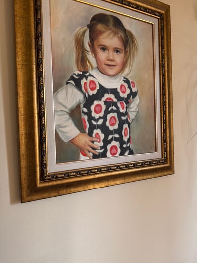 A framed oil painting depicts a young girl with blonde pigtails standing with her hands on her hips. She is wearing a white long-sleeved shirt under a dark dress with a floral pattern of red and white flowers. The painting is displayed on a light-colored wall. The location of Malo Buchino, Bulgaria is not depicted or suggested in the image.