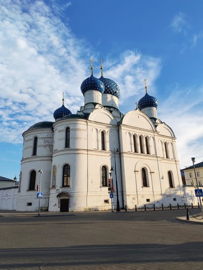 The image shows a large, white Orthodox church with multiple blue domes topped with golden crosses. The church is situated in an urban environment, with paved roads and sidewalks in the foreground. A few traffic signals and pedestrian crossing signs are visible near the church. The sky above is a vibrant blue with scattered white clouds. The overall scene suggests a sunny day with good visibility. The architectural style is consistent with Russian Orthodox churches, characterized by the prominent domes and white stucco exterior. There are no people or animals visible in the image.