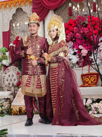 In the city of Meuliek, Indonesia, a newly married couple is pictured on their wedding day, adorned in traditional attire. The groom wears a deep maroon outfit with intricate gold embroidery and a matching headpiece. He holds a golden sash in his right hand. The bride is dressed in a complementary maroon gown, also embellished with gold thread and sequins, and wears a magnificent golden headdress adorned with white flowers. They stand side-by-side, facing forward, amidst a richly decorated backdrop of red and pink floral arrangements, draped curtains, and ornate chandeliers. To their left, a tufted white chair and a decorative white and gold structure are visible. The couple is standing on a white platform. The image captures a formal portrait of the couple, likely during a wedding ceremony or reception.
