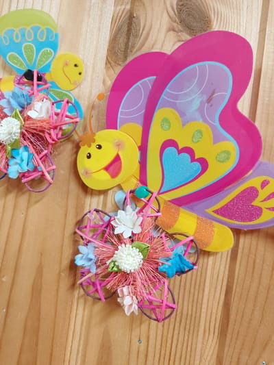 The image displays a close-up, overhead view of two colorful, craft butterfly decorations attached to a light-colored wooden surface, likely a wall or table. The butterflies are made of paper and other craft materials, featuring bright pink, yellow, and blue colors with glitter accents. One butterfly, on the right, is larger and more prominently featured, with elaborate wings and a striped yellow and orange body. The second, smaller butterfly, on the left, is partially obscured. Both butterflies are adorned with small, artificial flowers and have a rustic, woven circular element attached to their bodies, embellished with more tiny flowers and pink raffia. The wooden surface shows visible grain patterns and appears to be made of planks. There is no indication of the city of Alexandrovka, Russia, or any other specific location within the image.