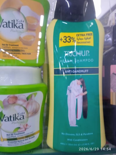 In the city of Ak-Suu, Kyrgyzstan, a retail setting is depicted. The foreground is dominated by two prominent hair care products. On the left is a green and white container of Vatika Naturals Hot Oil Treatment, featuring an image of garlic. To its right is a tall, green and yellow bottle of Trichup Herbal Shampoo, with a label stating "EXTRA FREE +33%," and an image of a person brushing long hair. The background is blurred, suggesting shelves lined with other products, typical of a store environment. The lighting appears to be artificial, casting a bright glow on the products. The overall scene is a static display of consumer goods.