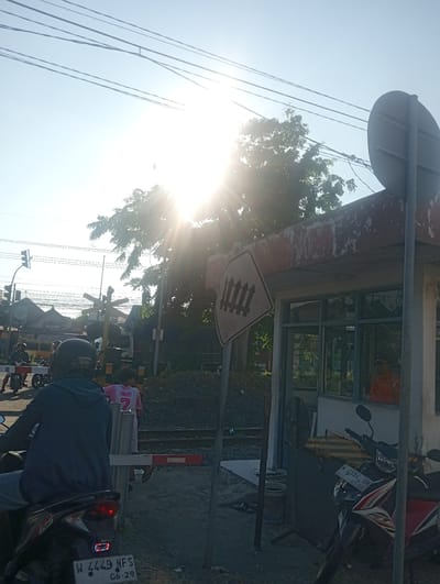 The image depicts a railway crossing in Tebel, Indonesia, under a bright, sunny sky with visible sun rays. In the foreground, a person on a motorcycle, wearing a dark hooded jacket and helmet, is facing away from the camera, waiting at the crossing. Behind them, a person in a pink shirt with the number "7" on it stands near the crossing gate.

The railway crossing features a yellow and white striped barrier gate, a railroad crossing sign with a fence icon, and traffic lights. Adjacent to the crossing is a small, open-air building with windows, possibly a ticket booth or control room, with a person in an orange shirt visible inside. Two motorcycles are parked in front of the building. Power lines are strung across the sky, and trees with lush green foliage are visible in the background. The ground appears to be a mixture of dirt and gravel. The overall scene suggests a typical day at a busy local transportation hub.