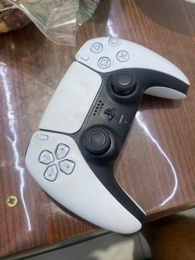 A white and black Playstation 5 controller is positioned on a dark wood surface, possibly a table. The controller is oriented diagonally, with the top right portion slightly further away from the viewer. The left side of the controller, closer to the viewer, displays the directional buttons, a microphone mute button, and a Playstation logo. The right side features the analog stick and face buttons: circle, cross, square, and triangle. A plastic bag containing what appears to be nuts is visible in the upper left corner of the image. The wood surface exhibits a visible grain pattern and some minor scuff marks. A single brass screw with a Phillips head is located to the left of the controller. The lighting suggests an indoor environment with a reflective glare across the surface. The context of Benkovski, Bulgaria is not evident from the image itself.