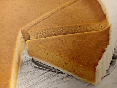 The image is a close-up shot of a pumpkin pie, partially sliced, resting on a white surface with a wood-grain pattern. The pie has a golden-brown filling and a white crust. Several slices have been cut, revealing the dense texture of the filling. The lighting is warm and appears to be artificial, casting subtle shadows. The image focuses on the texture and appearance of the pie, with no discernible background elements to suggest a specific location, and therefore, no connection to Rumaithiya, Kuwait can be made.