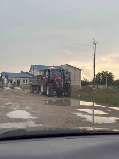 A red tractor with a trailer is parked on a gravelly, wet surface in front of a light-colored building with a blue roof in Husiatyn, Ukraine. There are puddles of water on the ground. A metal pole with a platform and ladder is visible in the background. The sky is overcast with a hint of sunlight.