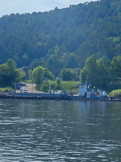 This image captures a ferryboat docked at a riverbank in Pristan', Russia. The ferry is a flat-bottomed vessel with a superstructure that houses the cabin and controls. It appears to be carrying vehicles, including a white van, a dark sedan, and a red truck, which are parked on the ferry. The ferry is positioned alongside a dirt ramp that leads up to a wooded area. A dense forest of pine trees covers a hillside in the background, with a clear sky overhead. The river in the foreground has a calm surface with ripples, reflecting the trees and sky. Two individuals can be seen on the deck of the ferry, one near the cabin and another closer to the stern. A sign on the ferry reads "ПАРОМ 94". The overall scene suggests a functional ferry service operating in a natural, forested environment.