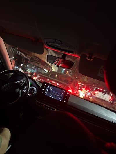 From inside a stopped vehicle at night, the scene reveals a traffic jam on a city street. The vehicle's dashboard, steering wheel with a Lincoln logo, and infotainment screen are visible. Through the windshield, multiple vehicles are clustered together, including a white ambulance with flashing red lights. A crosswalk is marked on the street. The mirror shows a vehicle behind and pedestrian in the road. Radio Station is set to 106.5. The location is believed to be Tizapan, Mexico.