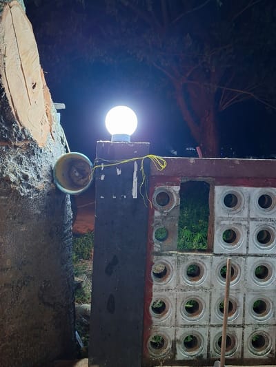In Meukokuthang, Indonesia, a dimly lit scene unfolds. To the left, a large tree trunk, freshly cut, stands prominent, revealing its textured bark and a clean, sawn surface. Attached to this trunk is a pale blue bucket. Adjacent to the tree trunk, a dark, rectangular post supports a bright, spherical lamp that casts a white glow. This post is attached to a section of a wall constructed from concrete blocks, each with a circular opening. The wall is topped with a weathered, reddish-brown frame. Through the gaps in the wall, green foliage is visible, suggesting an outdoor or semi-outdoor setting. The overall atmosphere is nocturnal, with the lamp being the primary light source in the immediate area. The ground is not clearly visible, but hints of dry vegetation and a tool with a wooden handle are present near the wall.