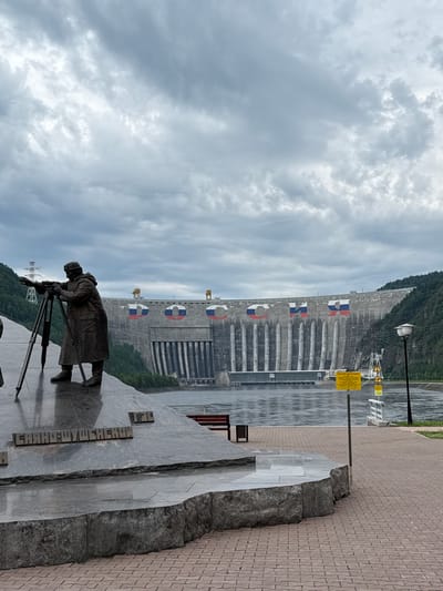 In Cheremushki, Russia, a monumental concrete dam dominates the background under a heavily clouded sky. The dam's facade is adorned with large, circular Russian flags. In the foreground, a bronze statue of a man in a long coat stands behind a surveying instrument on a tripod, appearing to be taking measurements. The statue is situated on a multi-tiered granite monument, inscribed with Cyrillic text that reads "SAYANO-SHUSHENSKOY." A river flows in front of the dam, and a paved walkway with red brick pavers leads to the right. Several yellow warning signs with triangular symbols are visible near the dam. The overall scene depicts a significant hydroelectric power facility and its surrounding commemorative area.