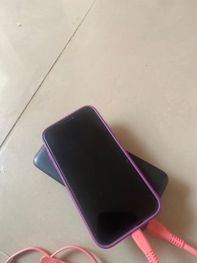 A dark-screened iPhone, enclosed in a purple case, rests on a black power bank. Two pink charging cables are plugged into the phone's charging port. The objects are situated on a tiled floor, with grout lines visible. The setting is the interior of a residential building in Wuro Leggal, Nigeria.