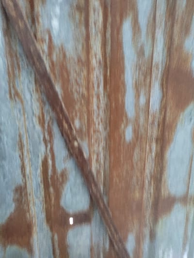 The image shows a close-up of a rusty corrugated metal surface. The metal is primarily a light gray color, with brown rust patches scattered across it. A thick, rusty metal rod runs diagonally across the surface, obscuring some of the rust patches. The image is most likely from a location near the coordinates 42.71, 75.232, which is in the city of Kegeti, Kyrgyzstan.