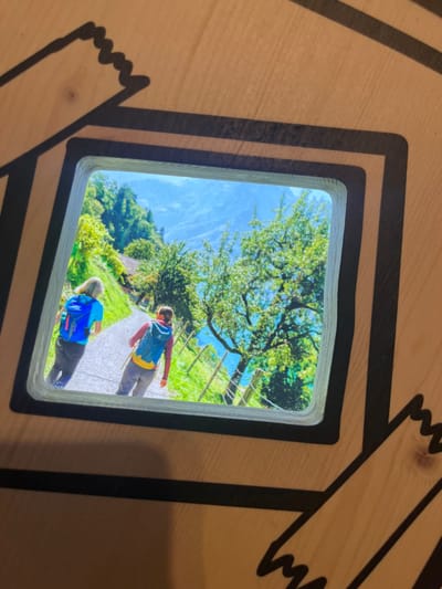 An image is presented within a smooth, rounded-corner rectangular cutout on a light-colored wooden panel. This display is located in Erstfeld, Switzerland. The wooden panel features bold, dark, irregular lines resembling stylized jagged edges, framing the central image and extending outwards. The displayed image depicts two individuals, viewed from behind, walking along a light-colored path in a vibrant, mountainous landscape. The person on the left wears a light blue short-sleeved top, dark pants, and a bright blue backpack with red accents. The person on the right wears a dark red long-sleeved top, light grey pants, and a light blue/teal backpack with yellow accents, appearing to hold a hiking pole in their right hand. The path is bordered on the right by a multi-strand wooden fence. Beyond the fence, a turquoise body of water, likely a lake, is visible at the base of steep, lush green hillsides covered in trees. A prominent green-leafed tree stands in the mid-ground to the right. In the distance, light blue mountains rise under a clear sky. Scattered buildings with reddish roofs are faintly visible on the distant slopes to the left of the path.