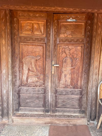 Two wooden doors intricately carved with relief panels. The top door features praying hands on the left, a figure in ornate clothing laying down in the middle panel, and a diamond-patterned panel on the right. The bottom door depicts a man working with an object on the middle panel with a diamond-patterned panel on the right and a solid panel on the left. An ornate border surrounds each panel.