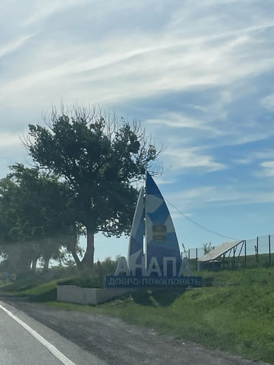 The image shows a sign at the entrance to the city of Anapa, Russia. The sign is shaped like a sailboat and has the text "АНАПА" (Anapa) in large blue letters. Below this, it says "ДОБРО ПОЖАЛОВАТЬ" (Welcome) in smaller blue letters. The sign is set against a backdrop of a blue sky with white clouds and green trees. In the background, there is a fence and what appears to be a solar panel. The foreground shows a paved road with a white line. The overall scene is a typical welcome sign for a city.