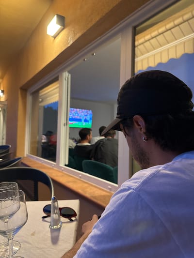The image shows a close-up, side view of a young man wearing a black baseball cap and a white t-shirt, facing away from the camera. He has short dark hair and a silver hoop earring in his visible ear. The man is indoors, possibly at a restaurant or cafe, given the table setting with wine glasses and sunglasses in the foreground. Through a large window behind him, another room is visible. In this inner room, a few people are seated, and a television screen displays a colorful image, possibly a sporting event or a video game. The time of day appears to be evening or dusk, as indicated by the sky visible through the window and the indoor lighting. The setting seems to be El Escorial, Spain, suggested by the provided location context. The overall mood is casual and relaxed.