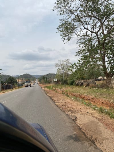 The image displays a two-lane asphalt road in Okengwe - Obehira, Nigeria, under an overcast sky with prominent grey clouds. The photograph is captured from a low vantage point, with a dark blue vehicle body panel visible in the bottom-left foreground.
The asphalt road, exhibiting minor surface imperfections and notable deterioration along its shoulders, particularly on the right, extends into the distance, curving slightly and ascending towards a series of hills. A light-colored heavy truck, possibly a dump truck, is observed in the left lane, moving away from the viewer. Two smaller vehicles are visible further ahead on the same road.
Along the right side of the road, a reddish-brown dirt shoulder transitions into a shallow, eroded ditch containing miscellaneous debris, including plastic bottles. Beyond this, the ground is covered with sparse dry grass and small patches of green vegetation. A large deciduous tree with green foliage occupies a significant portion of the right mid-ground. Further along the right, remnants of structures built with concrete blocks, appearing in disrepair or incomplete, are visible. Several tall, metal poles supporting solar panels and light fixtures (solar streetlights) are positioned intermittently along both sides of the road. A white, rectangular sign with unreadable text is mounted on one such pole on the right. Two dark-colored vehicles are parked off the road in the far distance on the right, near other structures.
The left side of the road features similar solar streetlights, scattered green vegetation, and distant buildings. The background is dominated by undulating, tree-covered hills. No human figures are discernible in the scene.