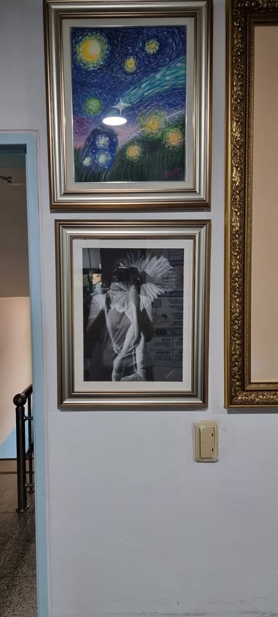 The image shows a section of a wall with three framed pictures hanging on it. The top picture has a gold ornate frame. The picture to the left is a reproduction of Van Gogh's "The Starry Night" with a silver frame. The picture to the right of "The Starry Night" is a black and white photograph of what appears to be a person in a ruffled collar and the silver frame. A light switch is located between the top picture and the right picture. The bottom of the image shows a portion of a doorway with a metal bar across it. The location is likely in Ucheon-myeon, South Korea, based on the file name or provided context, though this is not visually verifiable from the image itself.