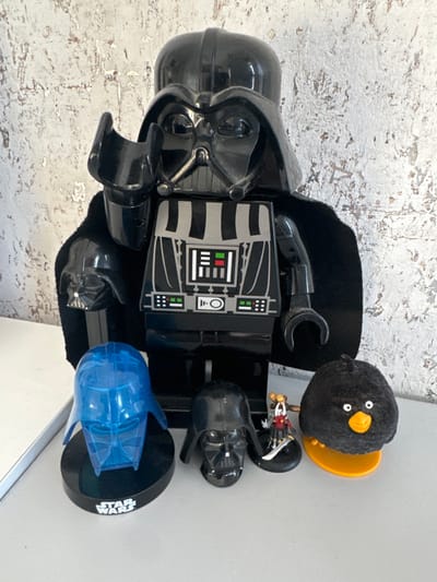 The image, taken in Matveevskoe, Russia, displays a collection of seven inanimate objects positioned on a flat, light grey surface against a textured background of light-colored, possibly peeling paint or plaster.
The central and largest item is a black Lego-style Darth Vader figure, approximately 30-40 cm tall, featuring a black fabric cape and a chest plate with printed grey, white, red, and green details. A smaller black Lego Darth Vader head is attached to the figure's left upper arm.
To the left of the large figure, a translucent blue plastic Darth Vader helmet rests on a black circular base labeled "STAR WARS" in white. Adjacent to this, a smaller, opaque black plastic Darth Vader helmet sits directly on the grey surface.
To the right of the large figure, a miniature figurine, approximately 5 cm tall, depicts a female character in red and gold attire, holding a silver sword, mounted on a black circular base. Next to it, on the far right, is a spherical black plush toy resembling a bird, with an orange beak and white eyes, situated on a yellow plastic clip-base.
No people are present, and no actions or interactions are depicted within the static arrangement of objects.