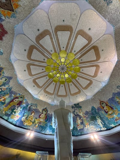 A white marble statue of a Buddha stands facing away from the camera, looking up at a domed ceiling. The ceiling is decorated with intricate mosaics depicting celestial beings and floral patterns. The mosaic on the dome is primarily white, beige, and gold, with a central pattern of radiating petal shapes. The surrounding wall is covered with a mosaic frieze of figures in colorful robes, some seated and some standing, set against a backdrop of blue and green landscapes. Two spotlights on the lower part of the wall cast bright beams upward, creating lens flare. The overall impression is of a serene and sacred interior space within a temple. This image was taken in the city of Chiang Khoeng, Thailand.