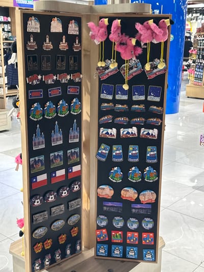 The image shows a display of souvenirs, likely in a shop, potentially located in Pudahuel, Chile, given the numerous Chilean-themed magnets and items. The display is divided into two levels inside a wooden frame. The top level features fridge magnets with images of landscapes, cities (Iquique, Santiago, San Pedro, Puerto Montt) and icons of Chile. There are also some flamingo plush keychains attached to the display. The bottom level of the display also features a variety of fridge magnets. These include magnets with images of Chilean landmarks, like a church in Chiloé, alongside magnets with the Chilean flag and items depicting footballs and wildlife. There are also magnets of the Chilean flag, and other general "I love Chile" motifs. The overall environment seems to be in a commercial area, with hints of retail goods in the blurry background.