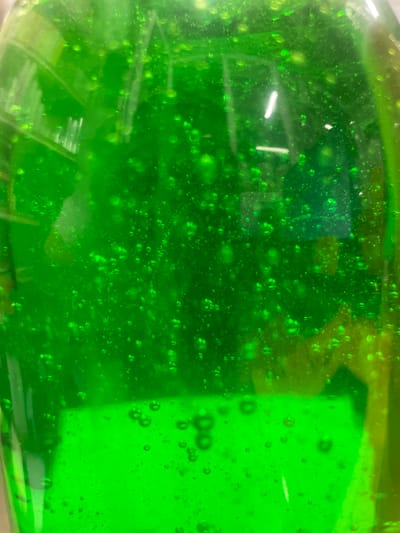 This is a close-up, eye-level, indoor shot of a vibrant green, viscous liquid, possibly hand sanitizer or a similar product, contained within a clear plastic bottle. The liquid is filled with numerous small, iridescent bubbles, some clustered together, creating a textured, almost effervescent appearance. Light reflects off the surface of the liquid and the bubbles, creating bright highlights. A distinct horizontal white line is visible in the upper right quadrant, likely a reflection from a light source. The lower portion of the image shows a flatter, more uniform layer of the green liquid with a slightly different texture, suggesting a separation or a change in the liquid's density. The background is a blurred, out-of-focus green hue, consistent with the liquid's color. The scene is devoid of any people or other identifiable objects, focusing solely on the visual properties of the liquid itself. There is no discernible connection to the city of Терволово, Russia, or any urban environment.