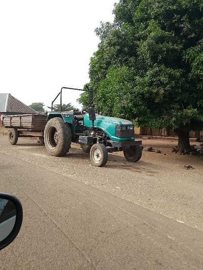 An teal-colored tractor with a brown, two-axle wooden trailer is positioned on the dirt shoulder adjacent to a paved road in Jibam, Nigeria. The tractor's large rear tires are soiled, and a black roll bar is visible above the operator's area. A large, densely leafed green tree occupies the right side of the frame, with bare earth and small rocks at its base. In the background, to the left, a building with a dark-colored corrugated metal roof is partially visible. The sky is bright white and overcast. A portion of a dark vehicle's side mirror is visible in the lower left corner, indicating the vantage point of the photographer. No individuals are visible.