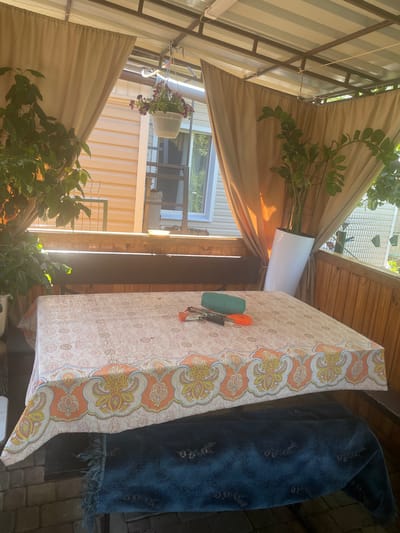The image depicts an outdoor covered patio area, likely in the city of Vysokyi, Ukraine. The scene is set with a table covered by a tablecloth featuring a patterned design of orange and yellow paisley-like motifs. On the table, several objects are visible, including a green cylindrical item, a pair of scissors, and what appears to be a keyring with multiple keys.
To the left of the table, a large potted plant with dark green leaves is situated, casting shadows on the tablecloth. A second plant, taller and also green, stands in a white cylindrical pot to the right of the table. In the background, a house with pale yellow siding is visible, with windows and a portion of a second story. The patio itself is constructed with wooden walls and a metal roof structure, supported by beams. Beige curtains hang from the roof, framing the view of the house. Beneath the table, a dark blue textured fabric, possibly a rug or blanket, is draped over a bench. The ground beneath the patio appears to be paved with bricks. The lighting suggests it is daytime, with sunlight filtering through the plants and curtains.