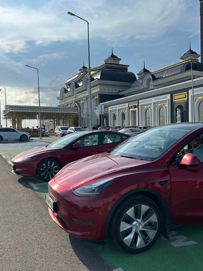 This detailed image captures an outdoor scene at the Kapitan Andreevo border crossing in Kapitan Andreevo, Bulgaria, featuring two red Tesla electric vehicles parked in the foreground, set against a backdrop of an ornate, large building under a partly cloudy sky.
In the immediate foreground, a red Tesla Model Y is partially visible on the right, showing its front passenger side, including the wheel, fender, and part of the hood and windshield. A person with dark hair and glasses is visible inside this Tesla, looking towards the right side of the frame through an open passenger window. Immediately behind and to the left of the Model Y, a red Tesla Model 3 is parked. Its front bumper displays a Bulgarian license plate, partially legible as "E 8899 AB." Both Teslas are parked on asphalt, within parking spaces marked with green paint.
In the middle ground, a grand, classically-styled building dominates the scene. It features multiple dark, domed roofs adorned with golden accents and spire-like finials. The building's facade is characterized by white and gray sections, further embellished with gold trim, ornate columns, arched windows, and decorative niches housing white statues, one of which is clearly visible on the right side of the facade. To the far left in the background, a modern, light-colored canopy structure with intricate metalwork is visible, extending over what appears to be another roadway or pedestrian area.
Several other vehicles are parked further back near the building, including a white SUV to the far left and various other cars indistinctly parked in the distance. The sky above is partly cloudy, with patches of blue sky and white clouds, and the lighting suggests either morning or late afternoon, with shadows extending from the vehicles and streetlights. Two tall streetlights are visible on the left side of the frame, and another is prominent in the upper center, partially obscuring the building.