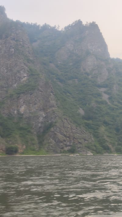 The image shows a wide river with rippling water in the foreground. In the background, a steep, rocky mountain covered in green vegetation rises from the riverbank. The sky is overcast and hazy. The scene appears to be a natural landscape. The location is identified as Ust'-Mana, Russia, suggesting a remote, possibly wilderness area. There are no people or man-made structures clearly visible in the image, implying a focus on the natural environment of the river and mountains.