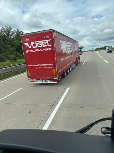 This image depicts a red semi-trailer truck with "VÖGEL SPEZIALTRANSPORTE" and "Fliegl" branding on its rear and side. The truck has the website "www.voegel-transporte.com" and the slogan "Eine starke Familie seit 1961." printed on the back. The license plate is visible as "BZ 230 FK". The truck is on a multi-lane highway, likely the A4, given the context of Klipphausen, Germany, and its proximity to Dresden and Chemnitz. The sky is cloudy with some patches of blue. Other vehicles are present on the highway in the distance. The photo appears to be taken from another vehicle traveling on the highway, with the edge of a windshield wiper visible in the foreground. The surrounding environment consists of green fields and trees.
