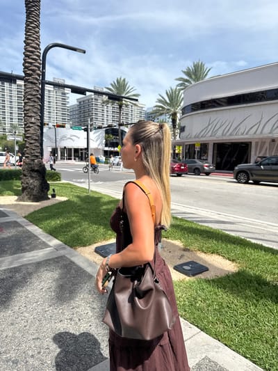 This image depicts an outdoor urban street scene in Bal Harbour Village, Florida, United States, specifically at or near Harding Avenue. The scene is illuminated by bright sunlight under a clear blue sky with scattered clouds.
In the foreground, a female subject is captured from the mid-back and right profile, standing on a paved sidewalk. She has long, blonde hair styled in a high ponytail and wears a dark brown sleeveless, possibly maxi, dress. A dark brown leather bucket bag with a tan shoulder strap is carried over her right shoulder. A silver-toned watch is visible on her left wrist. She is looking towards the left side of the frame. To her immediate left is a large palm tree trunk and a black street light pole.
Across a grassy median and a multi-lane road, several vehicles are present, including a red sedan, a grey sedan, and a dark SUV. On the opposite side of the road, a prominent white commercial building with a distinctive curved facade and horizontal grey accent lines is visible on the right. Further down the street to the left, another white building with a rounded entrance is present near an intersection where a "Harding Ave" street sign is legible. Multiple pedestrians are observed in the background, walking or crossing the street, with one individual seen on a bicycle. The distant background features several tall, multi-story buildings, characteristic of luxury condominiums or hotels, interspersed with numerous palm trees.