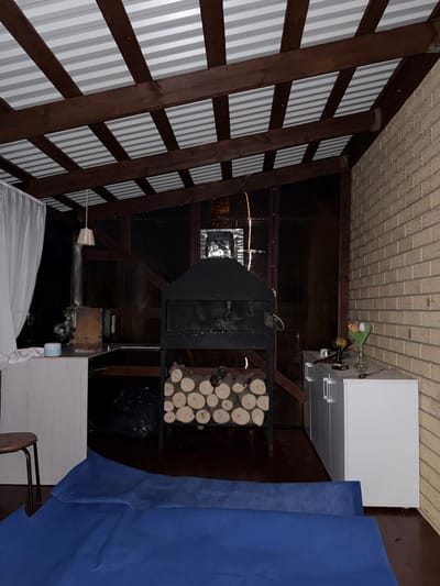 This media file captures an indoor scene, likely a covered porch or outdoor living space, possibly in Столбище, Russia. The perspective is rotated, making the ceiling appear as a floor and vice-versa.
The scene is furnished with several objects. To the left, a slatted wooden structure, possibly a wall or screen, is visible. In the center, a large black barbecue or smoker sits on a metal stand, with a stack of cut firewood neatly arranged beside it. To the right, a white cabinet with drawers is positioned, and a large piece of blue fabric, possibly a tarp or curtain, is draped alongside it. Further to the right, a simple chair with a wooden seat and metal frame is partially visible.
The lighting suggests it is nighttime or very dim. The overall impression is one of a functional outdoor space, perhaps set up for cooking or entertaining. There are no people visible in the frame. Notable details include the texture of the brick-like ceiling material, the distinct pattern of the slatted wall, and the arrangement of the firewood. The presence of what appears to be a plant in a vase on top of the white cabinet, along with a small tool, suggests recent activity or preparation.