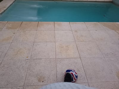 A pale tiled patio surrounds a swimming pool filled with blue water. The tiles are rough, with variations in color. A striped ball sits on the tiles near the bottom edge of the frame. A person's leg is partially visible at the very bottom of the image, close to the ball.