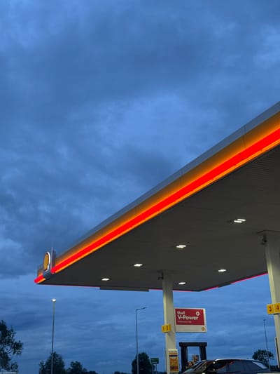 The image shows a Shell gas station canopy with a red and yellow neon sign in Pocklington, UK. The sky is blue with a few white clouds and it appears to be dusk. There are streetlights on either side of the canopy. There are multiple lights on the underside of the canopy. A sign for Shell V-Power fuel is visible on the right. There is a dark-colored car parked at the pump under the canopy.